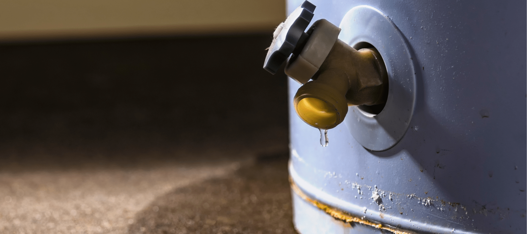 Close-up image of a weathered blue water container with a yellow plastic tap, slightly dripping, set against a dark background with sunlight highlighting the droplet and rust around the base