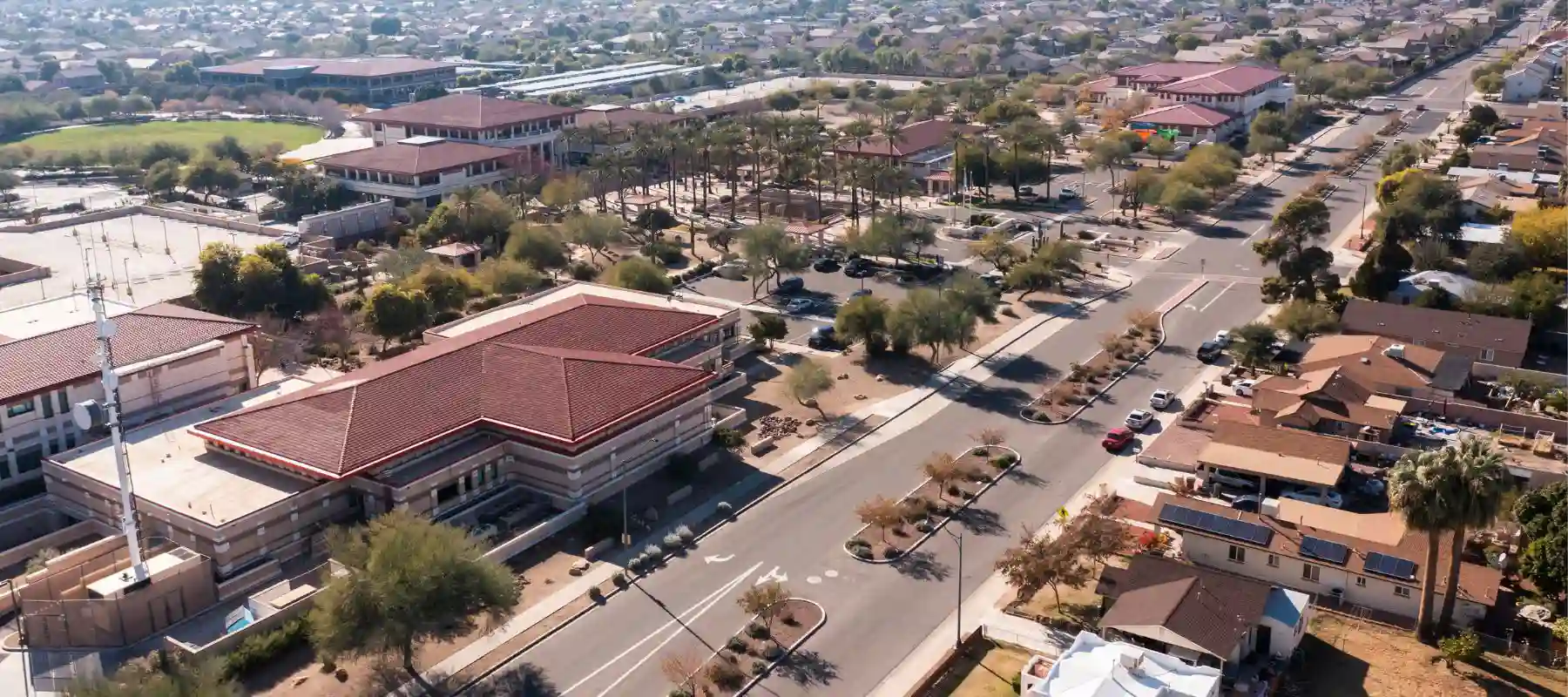 Aerial view of a suburban area featuring a prominent building with a red-tiled roof at the intersection, surrounded by single-story houses and tree-lined streets, on a clear day with sparse traffic