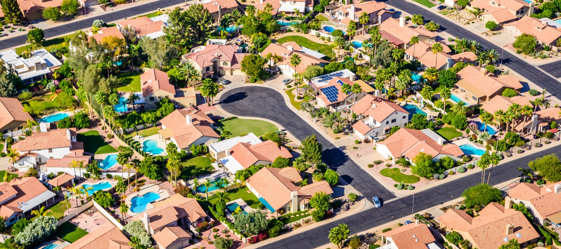 Bird's-eye view of a residential neighborhood with rows of houses featuring red-tiled roofs and landscaped yards, several with swimming pools, along curving streets on a sunny day