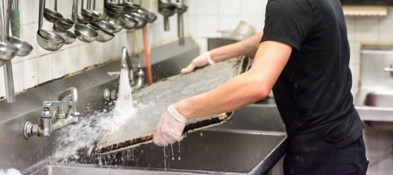 A person wearing white gloves rinses a large, rectangular baking sheet under running water in a commercial kitchen sink, with ladles hanging above